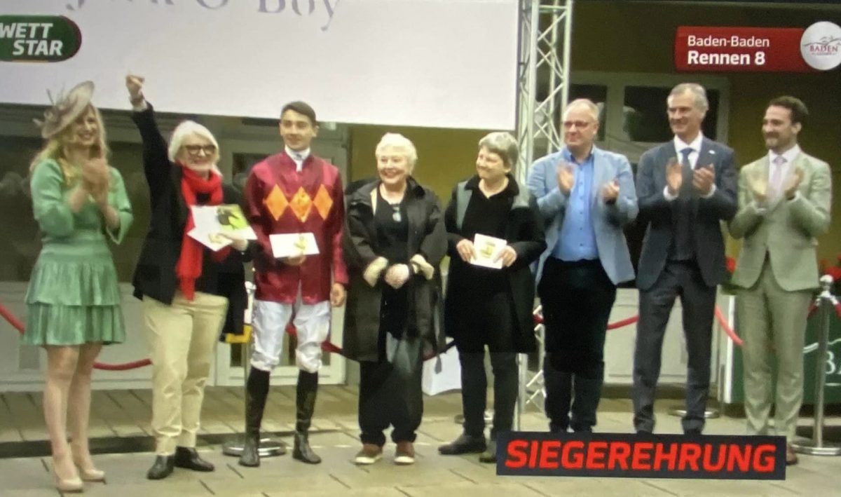 Susanne Born, owner of Jack O'Boy, raising her hand high in triumph at the trophy presentation. 
To her right, jockey Hugo Boutin, Manuella Line Groll, trainer Gina Rarick and racecourse representatives.